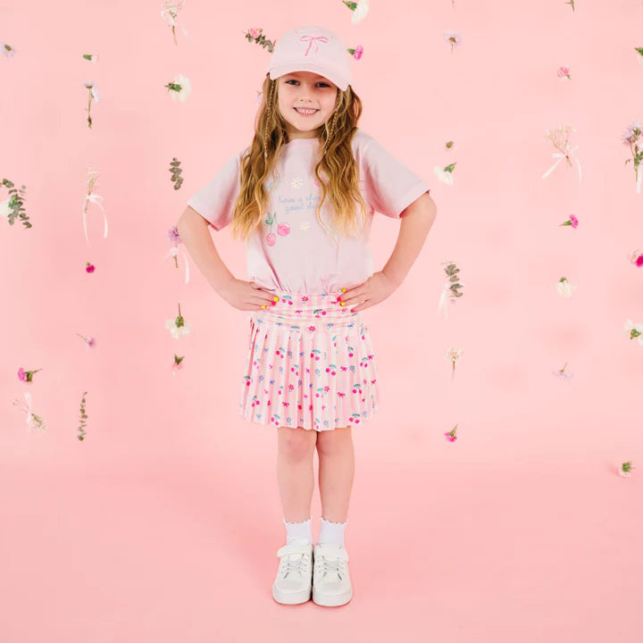 Model wearing Sweet Wink Ballet Cherry Good Day short sleeve T-shirt with cherry and bow patch, paired with athletic skort.