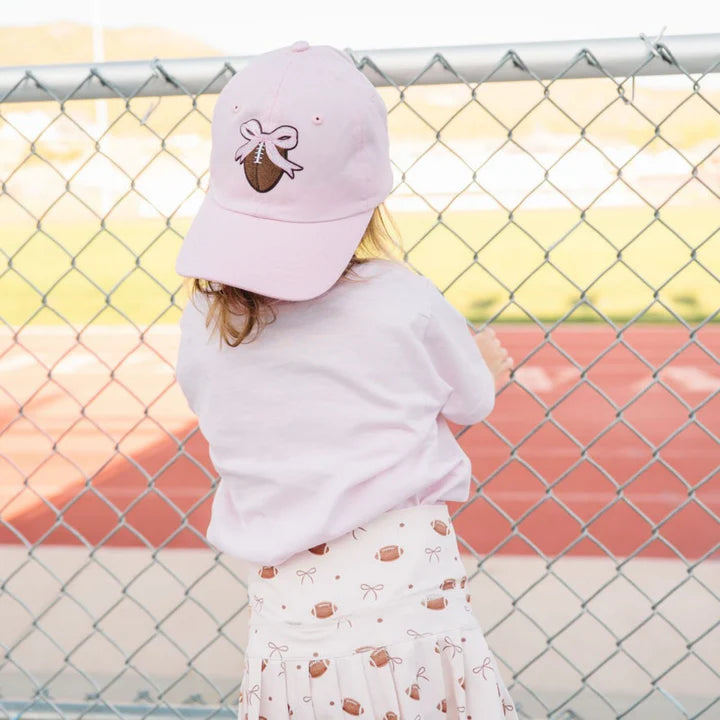 Model wearing Sweet Wink Game Day Athletic Skort paired with a football-themed baseball hat and short-sleeve t-shirt for a sporty look.