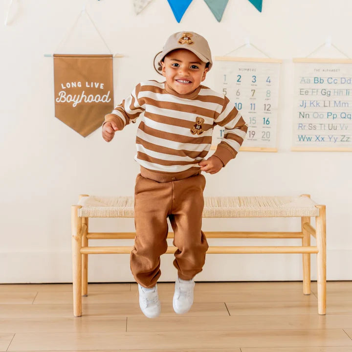 Model wearing Sweet Wink brown joggers with teddy bear patch, striped sweatshirt, and baseball hat, showcasing casual, stylish outfit.